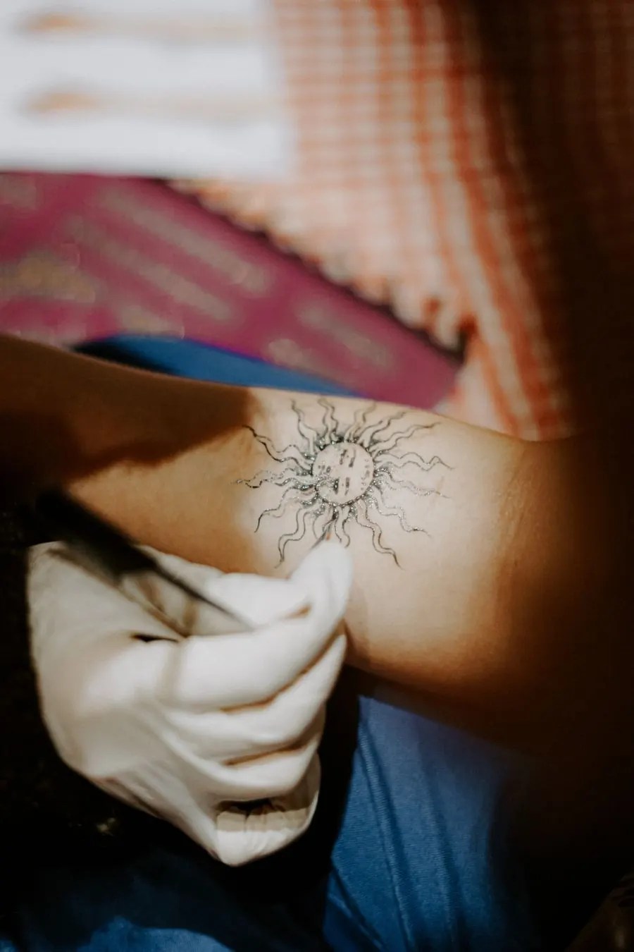 Tatoueuse gantée traçant un petit soleil au trait fin sur une peau claire