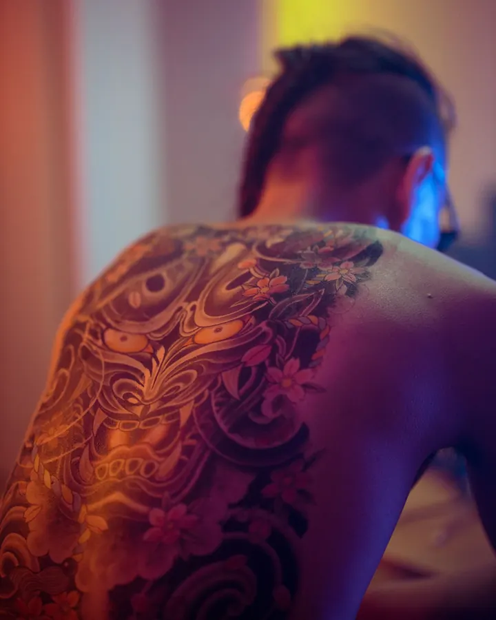 Dos tatoué d'un grand visage de démon et de fleurs, éclairé par une lumière orangée.