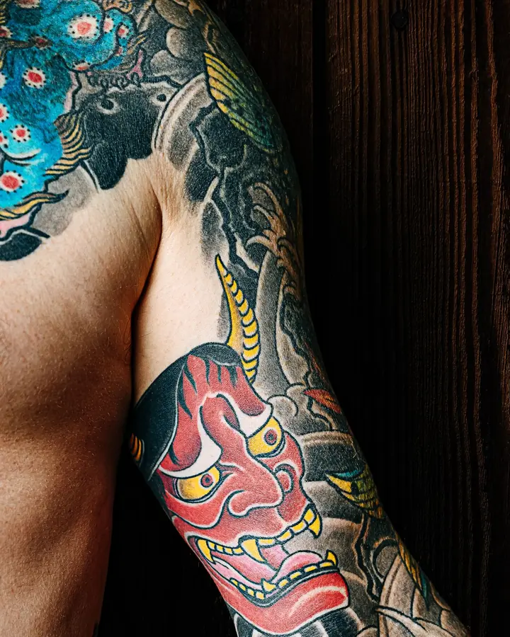 Épaule tatouée d'un lion gardien bleu, qui descend vers un masque hannya rouge sur le bras.