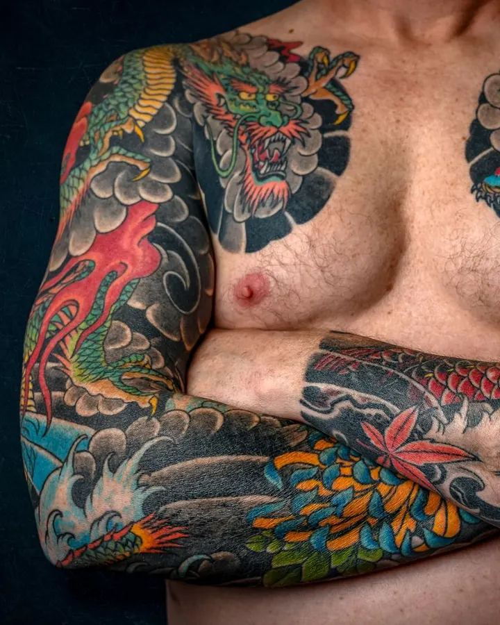 Épaule et bras tatoués d'un dragon vert aux flammes rouges, avec un fond de nuages noirs.