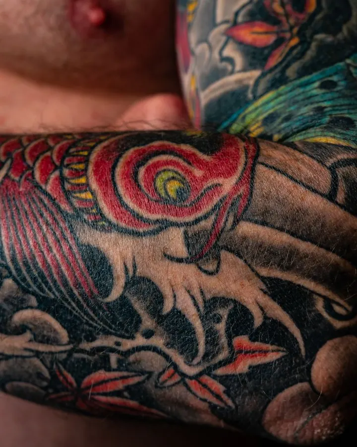 Avant-bras tatoué d'une carpe koï rouge qui remonte des vagues noires, parmi des feuilles d'érable.