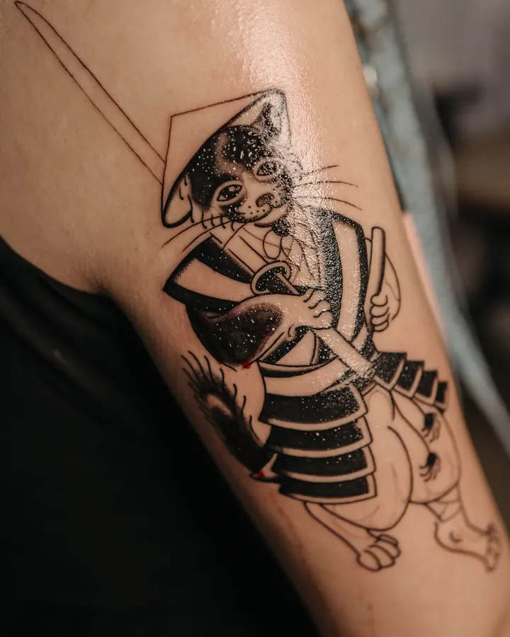 Tatouage en noir sur le bras : un chat en armure de samouraï portant un chapeau conique.